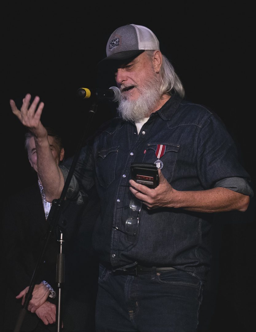 An man with grey hair and a grey beard, wearing a denim shirt, jeans and a ball cap, speaks before a microphone.