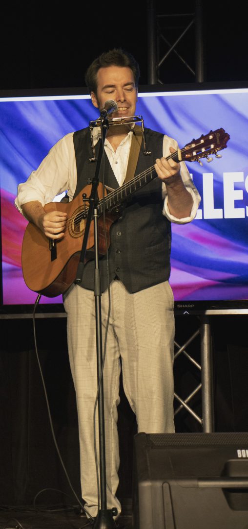 A man wearing a dark vest, white shirt and beige pants holds a guitar while he sings into a microphone.