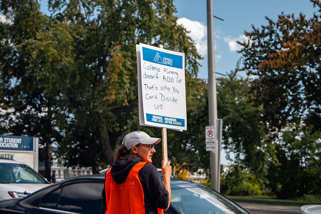 A striker holds a sign that says "College funding doesn&squot;t ADD up. That&squot;s why you can&squot;t divide us."