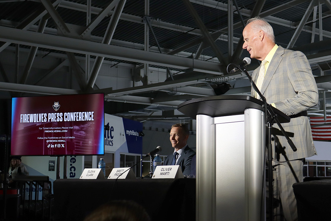 A man in a grey suit stands at a podium in front of a microphone.