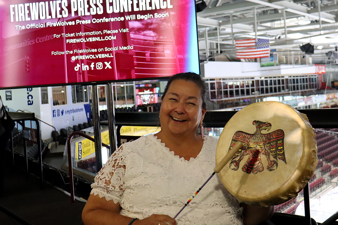 An Indigenous woman in a white blouse holds a drum and drumstick.