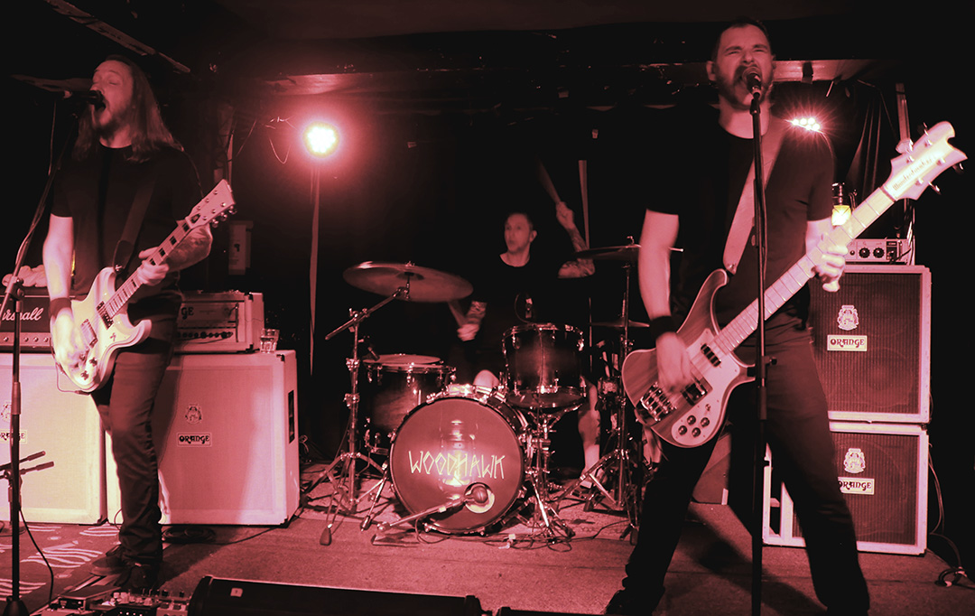 Three members form the rock trio Woodhawk seen on stage performing guitars, drums, bass, and vocals