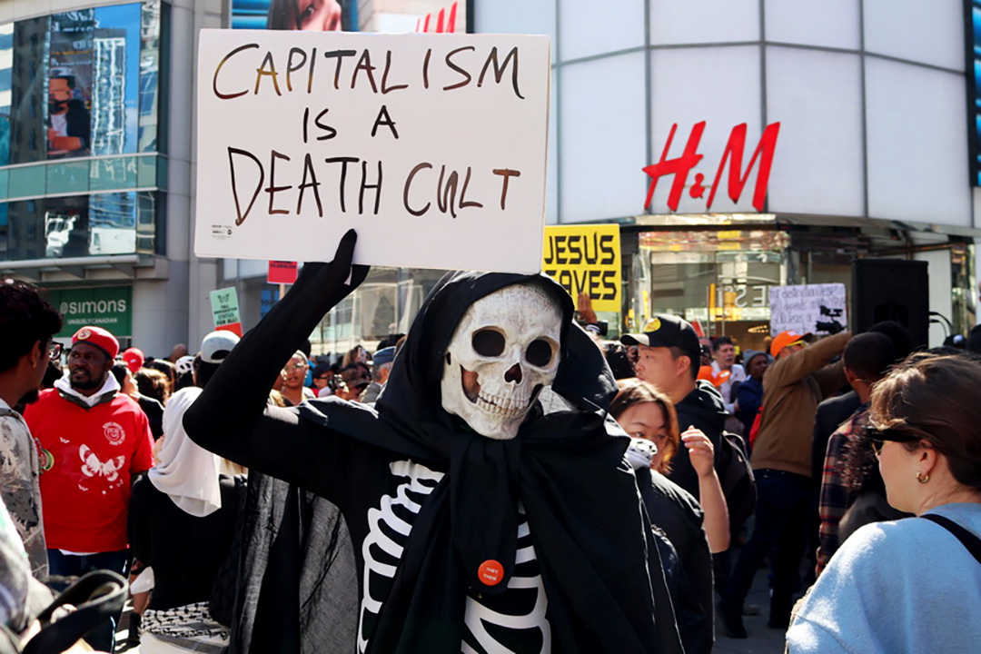 Person in a black and white skeleton costume holding a sign up that says "Capitalism is a Death Cult."