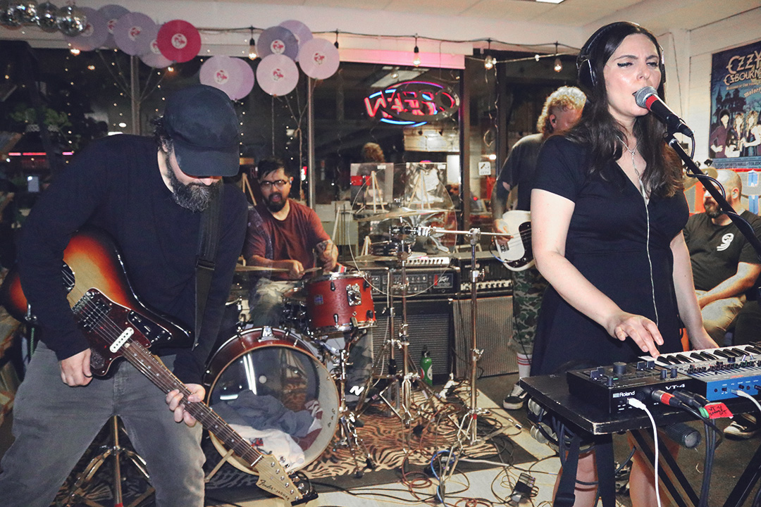A woman plays a sampler keyboard while singing into a microphone. A man wearing a cap plays guitar and a man in a red shirt plays the drums behind them.