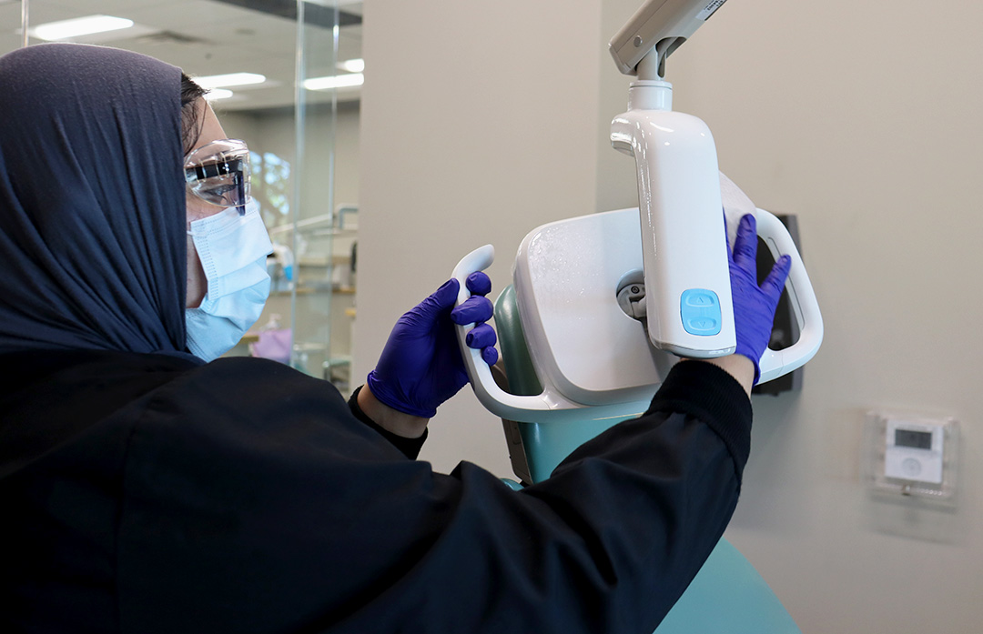 A woman in black medical scrubs wearing a dark hajib and purple gloves sanitizes a light.