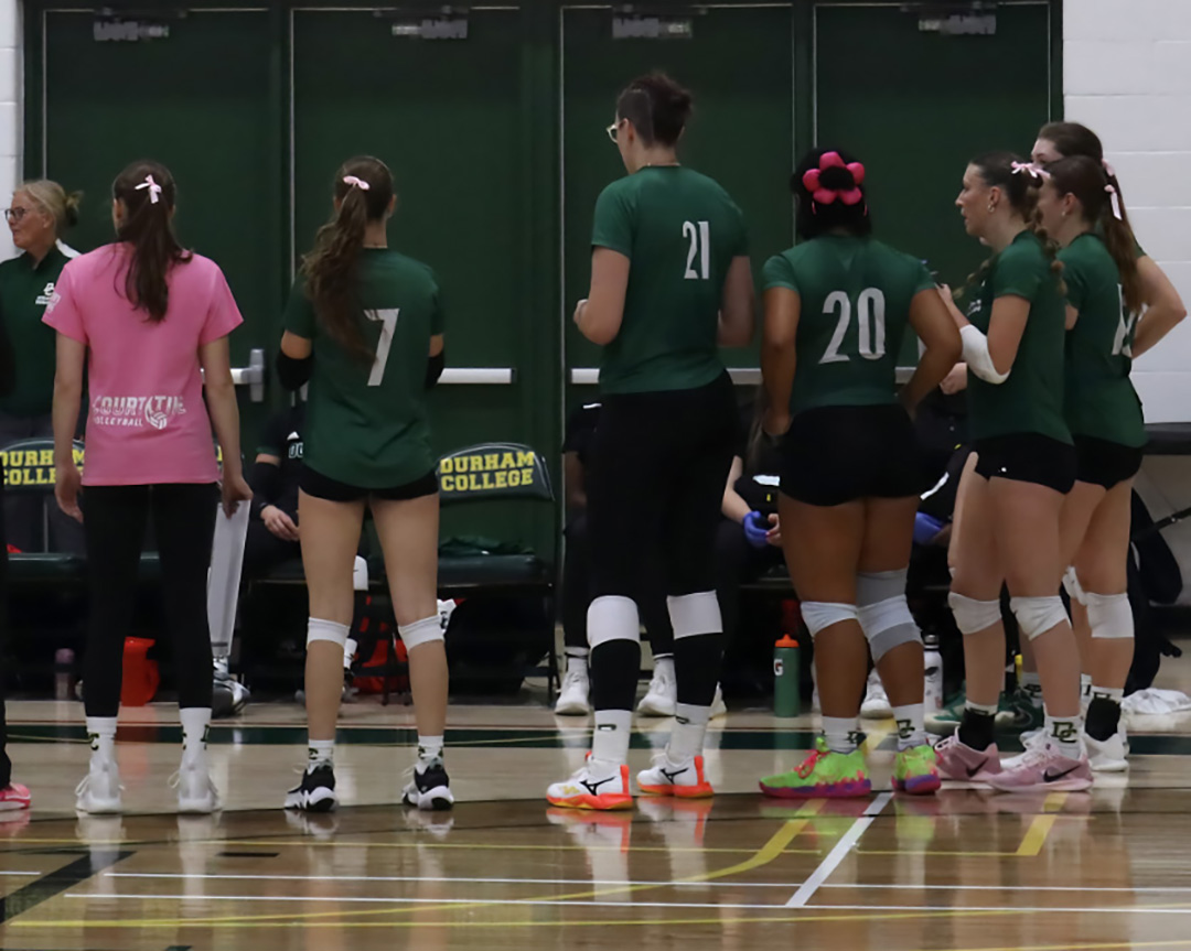 A group of Durham Lords Volleyball players hudled wearing their green jerseys and white bows. One girl is wearing the "Think Pink" T-shirts that the team wore during warm-up.