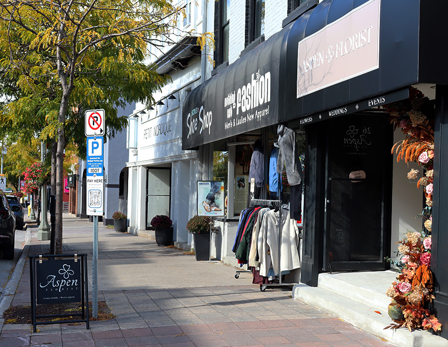 A sidewalk in front of several businesses in downtown Bowmanville. The first store is painted black, except for the words "Aspen Florist" which is written in white with a pink rectangle border.