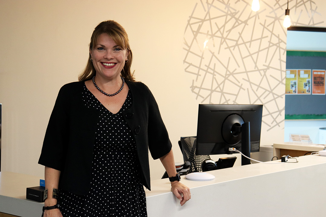 Women dressed in a black dress with white polkadots and a black cardigan that goes to her elbows. She stands in front of the reception desk of an office space.