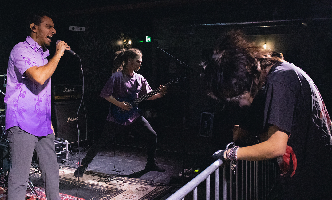 A young person to the right of the frame is headbanging and holding a guardrail. To his left, there are two men performing on stage.