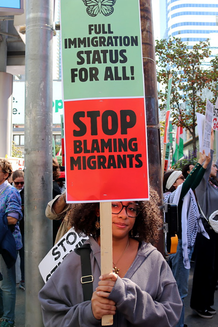 Young female adult with curly hair, partially covered by a sign she is holding that says "Full Immigration Status for All" and "Stop Blaming Migrants."
