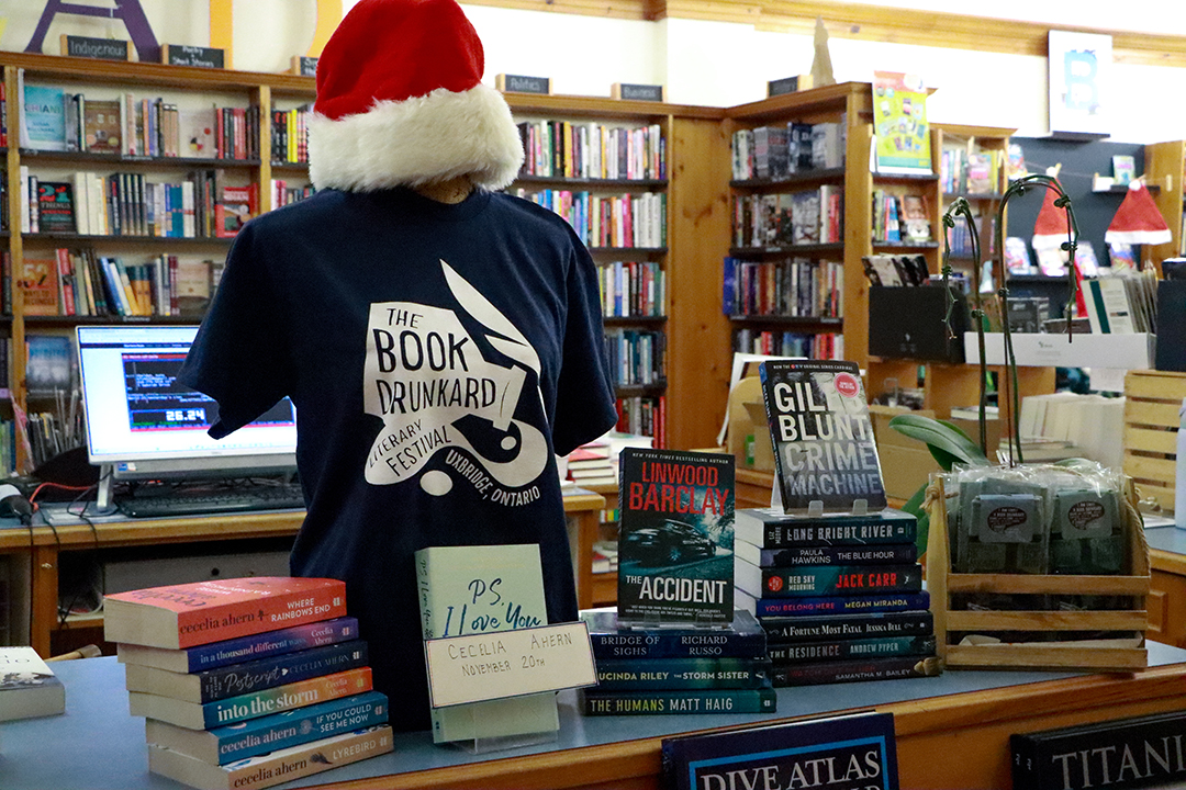 A bookstore display features a mannequin wearing a navy Book Drunkard Festival T-shirt and a red-and-white Santa hat. Surrounding it are stacks of novels, including Linwood Barclay’s The Accident, and several shelves filled with books in the background.
