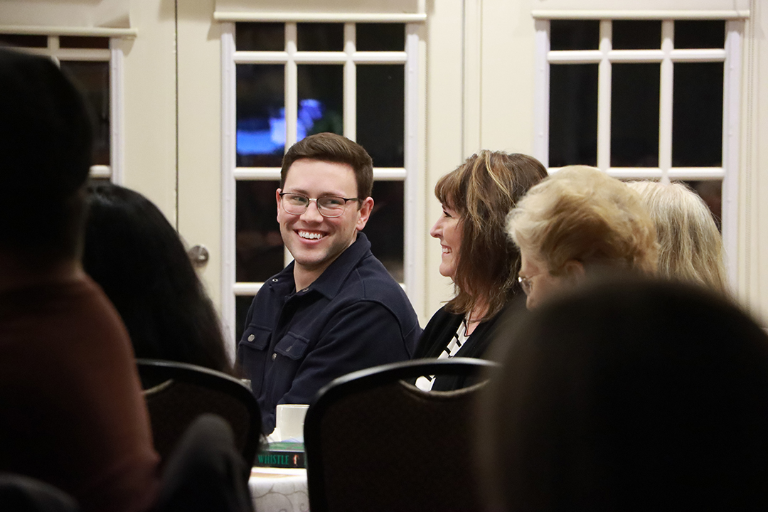 A young man wearing glasses smiles at a female attendee while seated at a round table. Window panes are visible behind the group, and several people around him are turned toward the stage.