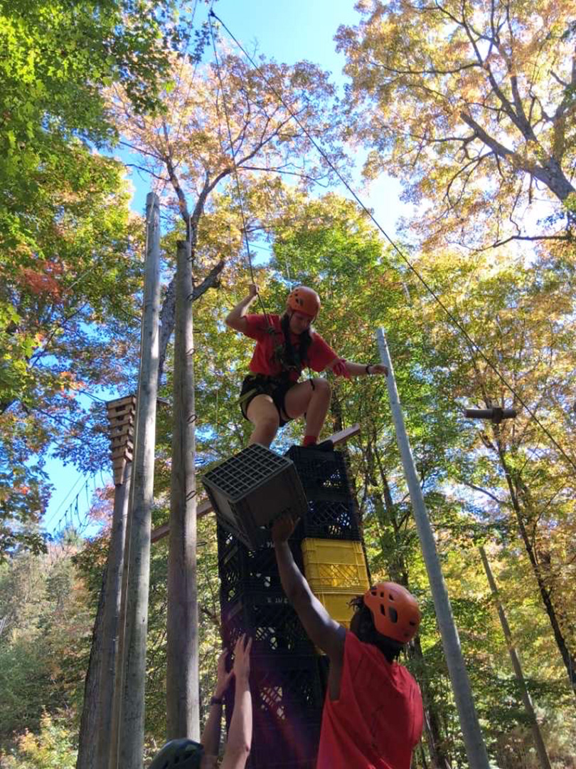 A girl stands on top of a stack of milk crates in a forest. Her team stabilizes the crates and passes them up to her one at a time.