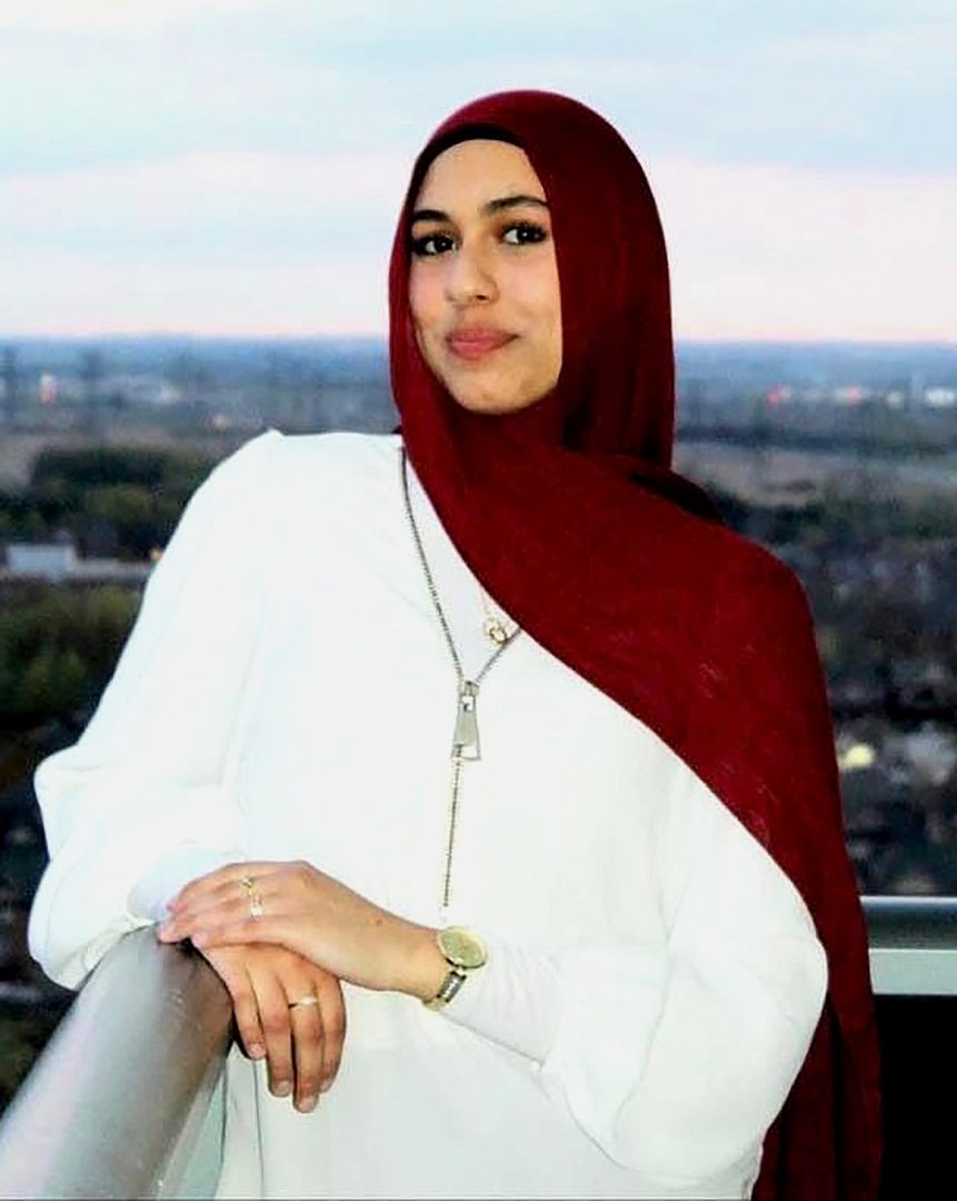 A young woman wearing a white dress and red hijab looks at the camera. Her arm is resting on a railing with a blurred urban backdrop behind her