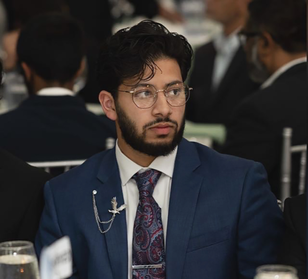 A young man wearing glasses and a formal deep blue suit looks towards the right, seated in a room with people in the background