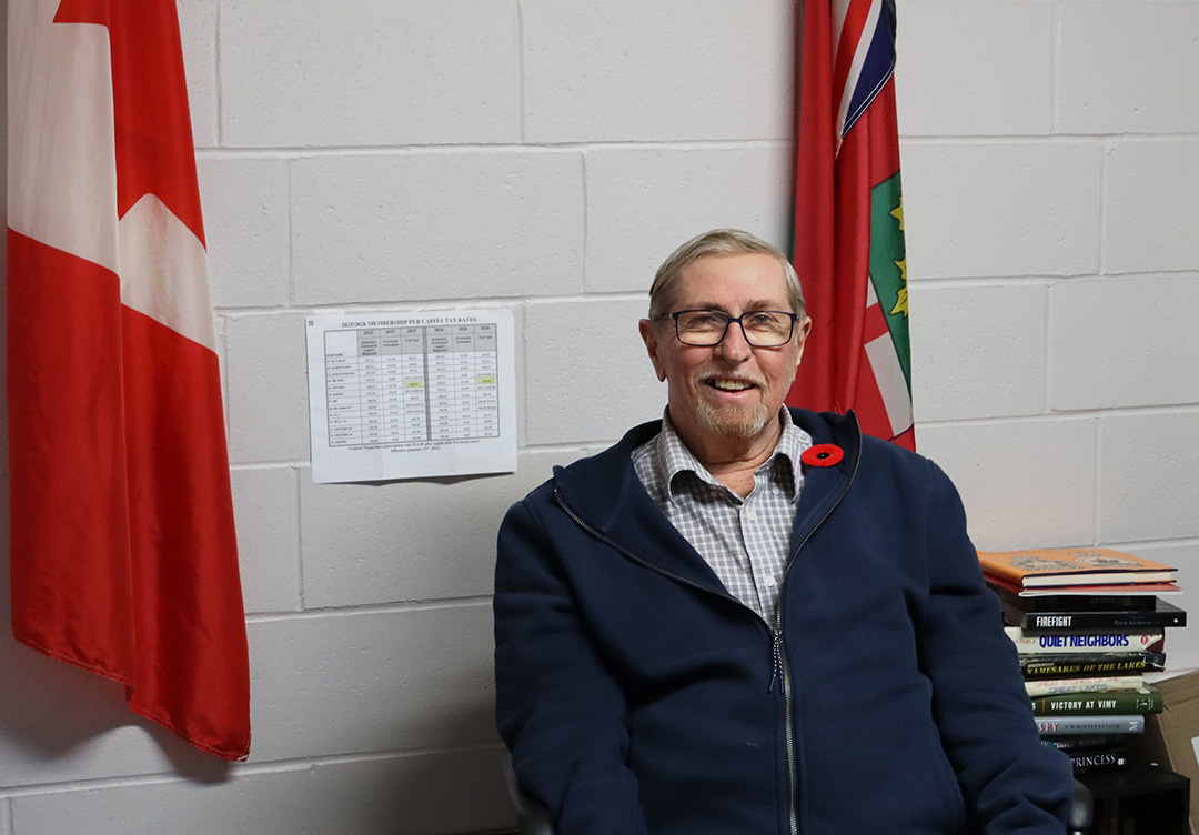 A man sitting in a chair wearing a dark blue zip up hoodie, a blue and white collared shirt underneath a a red poppy pinned over his heart. Behind him are two hanging flags, to the left a Canadian flag and to the right an Ontario flag.