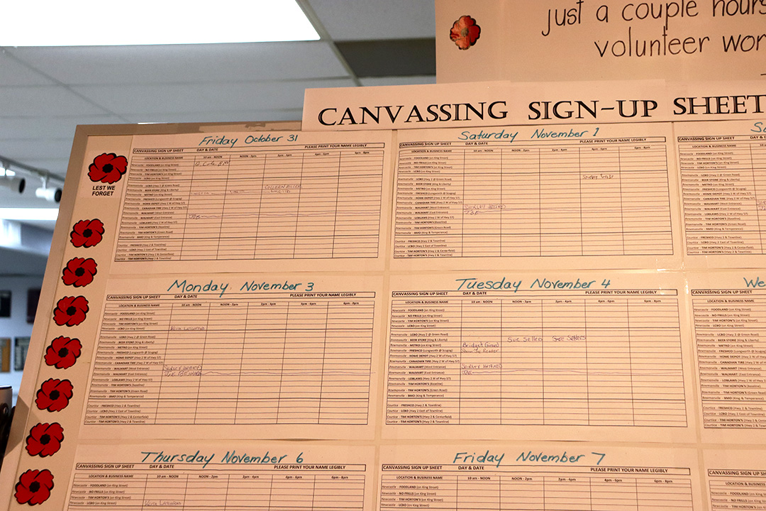 A large board with dates, locations and times for canvassing local businesses to sell poppies. The top of the board reads "Canvassing Sign-up Sheet." The board is lined with red poppy stickers.