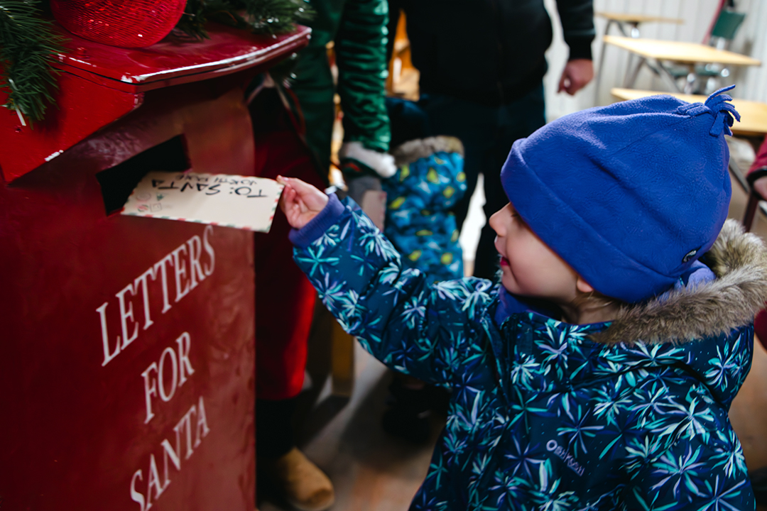A small child wearing a blue winter hat and patterned snow jacket reaches up to place a handwritten letter addressed “To: Santa” into a large red metal mailbox labelled “LETTERS FOR SANTA.” The child is smiling slightly as adults and another child are bundled in winter clothing standing in the background.