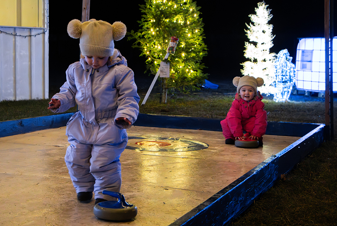 Two young girls bundled in winter snowsuits play on a small curling lane illuminated by holiday lights. One child in a lilac snowsuit walks forward while pushing a blue curling stone, while the other child in a bright pink snowsuit sits behind her holding a red stone and smiling. Christmas trees decorated with warm lights glow in the background.