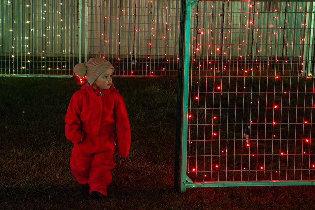 A small girl in a bright fuchsia snowsuit and beige knit hat walks through a fenced light maze at night. Red and warm white string lights hang from metal panels around her, casting a colourful glow on the grass. She looks to her right as she passes through.