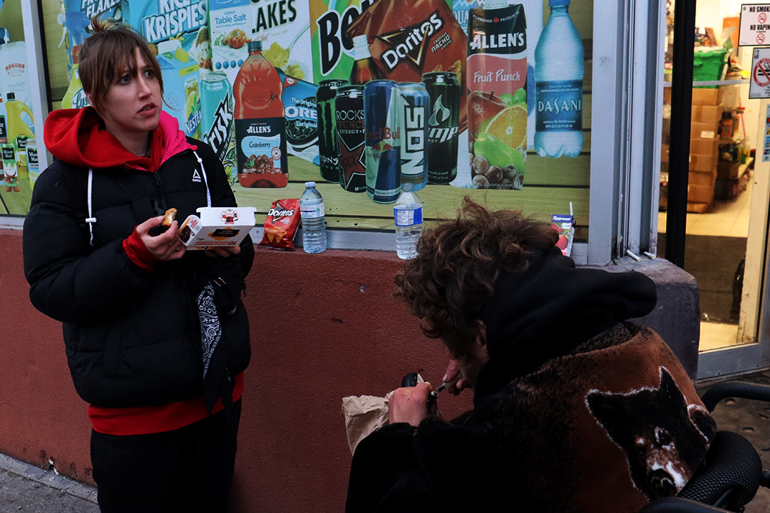 A young woman wearing a red hoodie and black jacket eats chicken nuggets. A man in a wheelchair faces her.