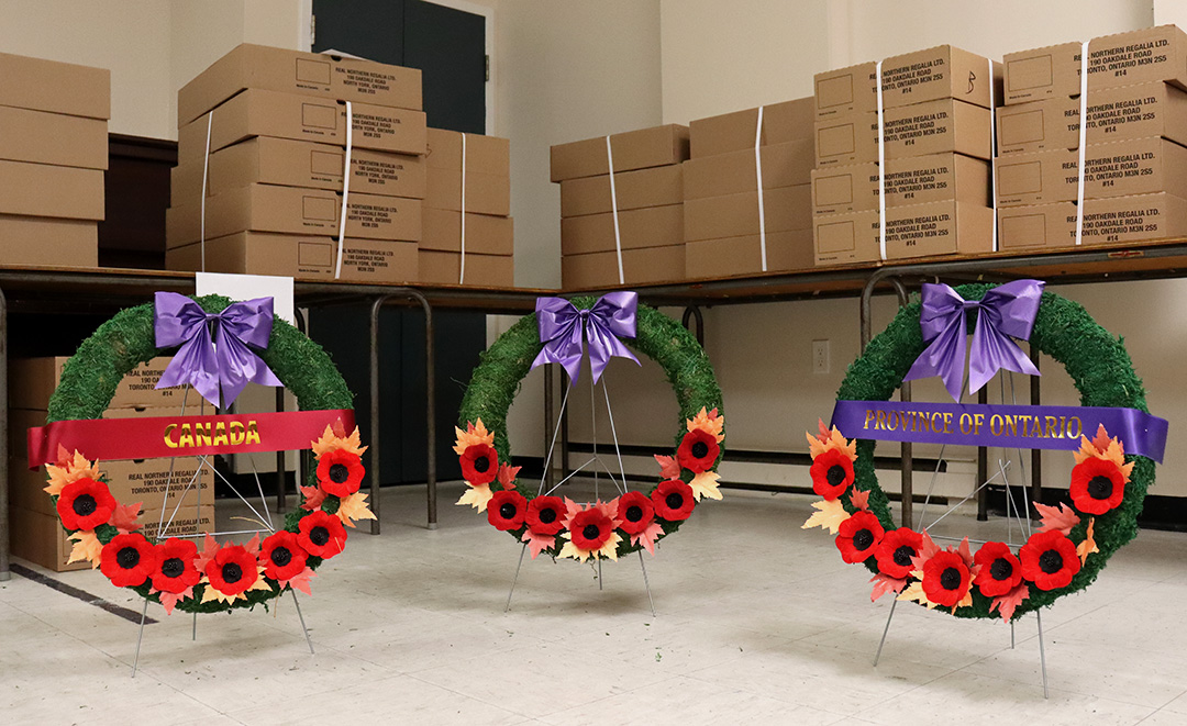 Three green wreathes with red poppy&squot;s decorated the bottom half and a purple bow tied at the top. The left wreath has a ribbon that reads "Canada" while the right wreath has a ribbon that reads "Province of Ontario," the middle wreath has no ribbon. Behind the wreathes are tables stacked with brown cardboard boxes.