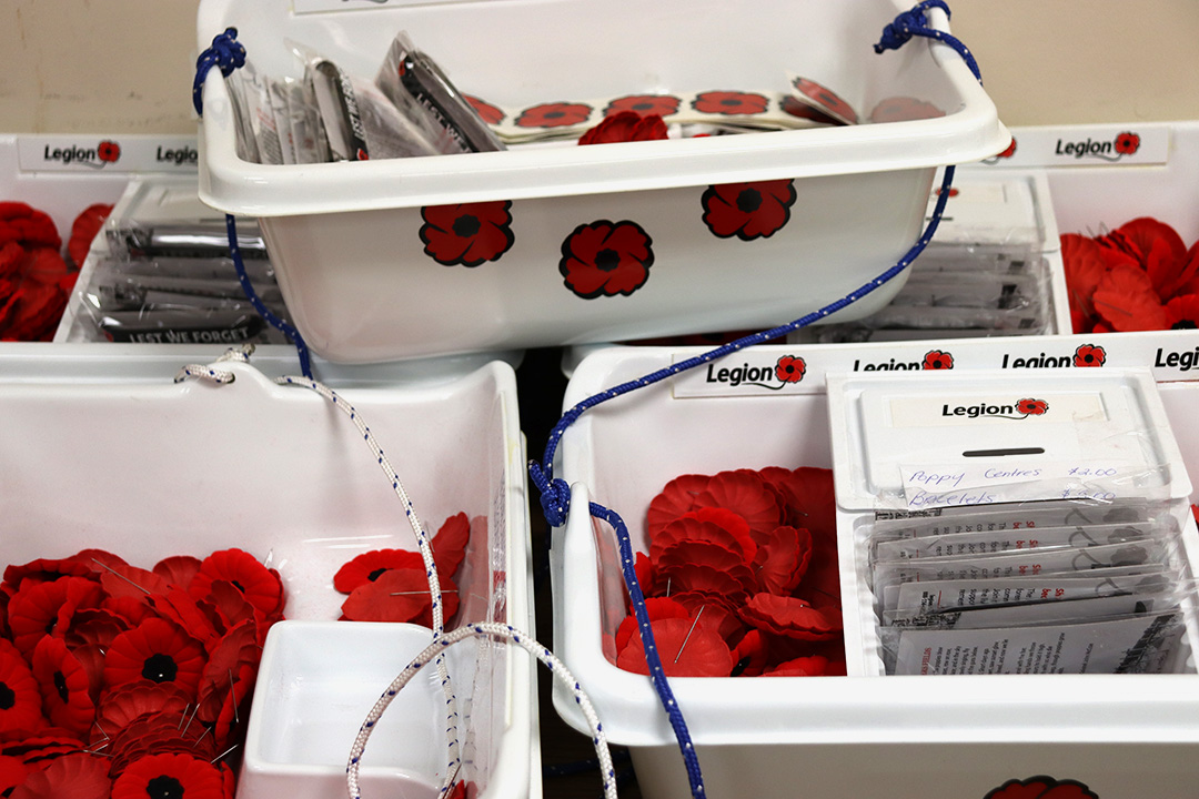 Several white bins that are full of red poppies and rubber bracelets that read, "Lest we forget." The bins are stacked on top of each other. Each bin has blue or white rope tied to the left and right sides to make them easy to carry.