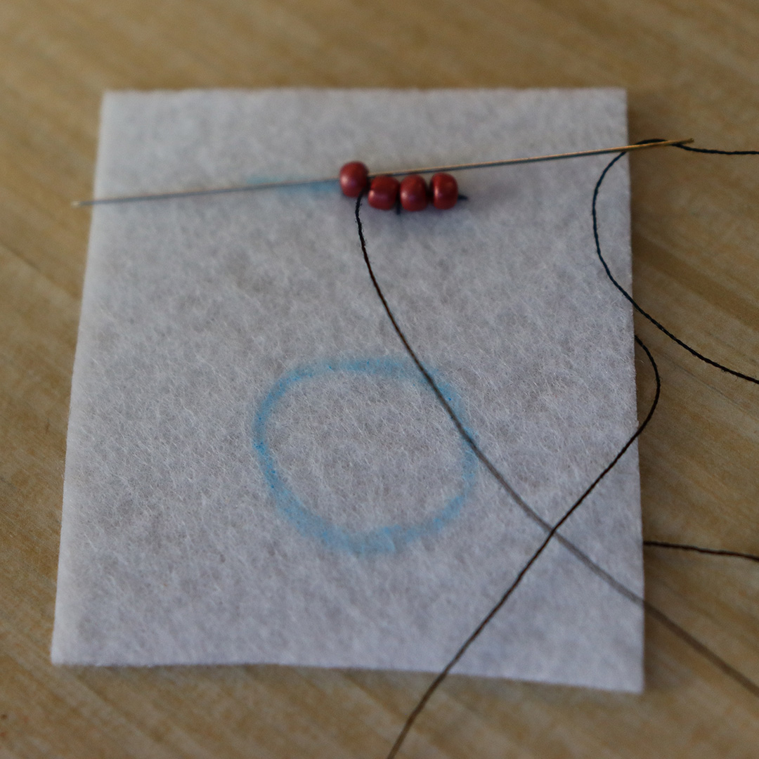 A piece of white felt with a needle, thread and red beads on top.