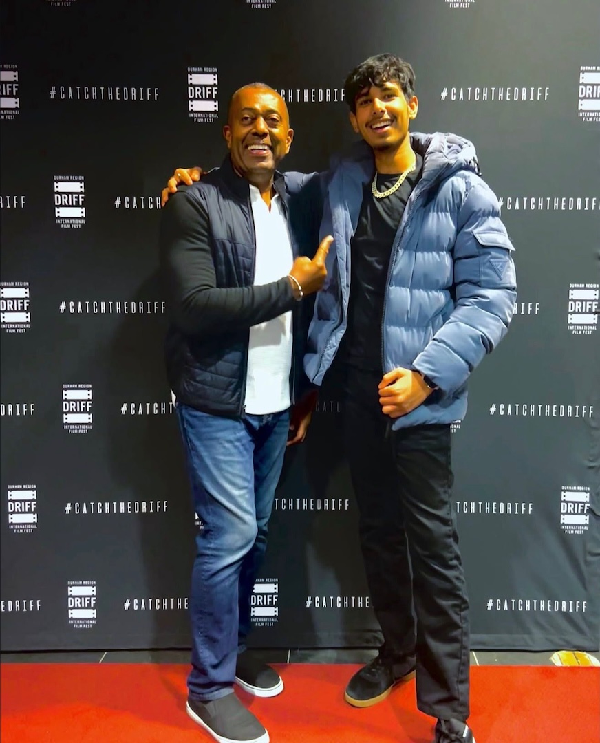 Two men pose against a black background. The man on the left is wearing a white t-shirt and a black jacket and is pointing to his left. The man on the right is in a blue jacket and smiling.