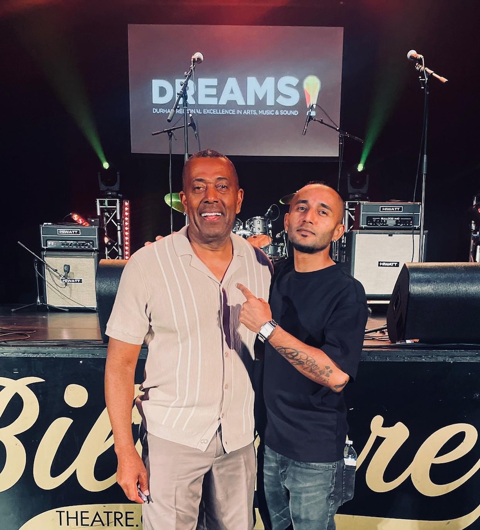 Two men stand in front of a stage with amplifiers and a screen in the background. The man on the left is smiling and wearing a beige shirt, and the man on the right is pointing to his right and wearing a black t-shirt.