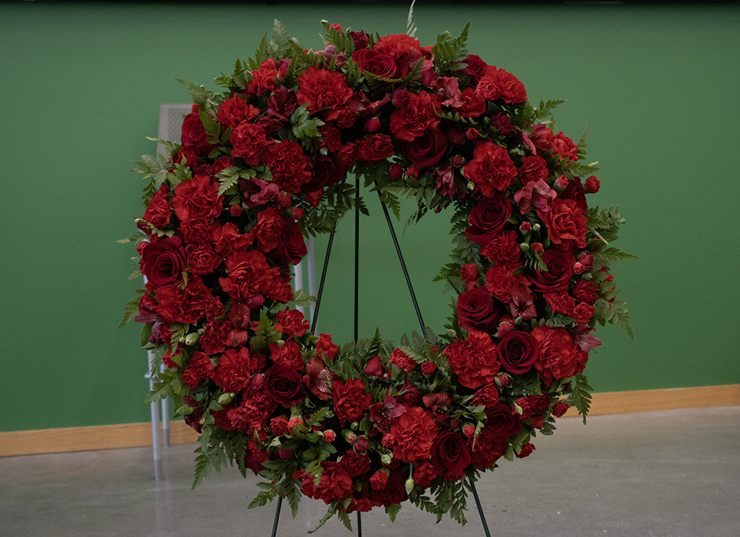 A wreath of red roses sits on an easel in front of a green wall.