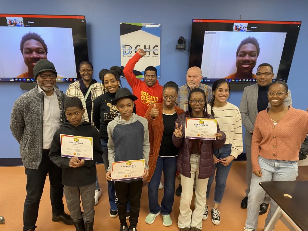A group of children and adults pose in a classroom. Three children hold certificates, and a man appears on a screen behind them via video call. Staff members and parents stand behind the children, smiling.