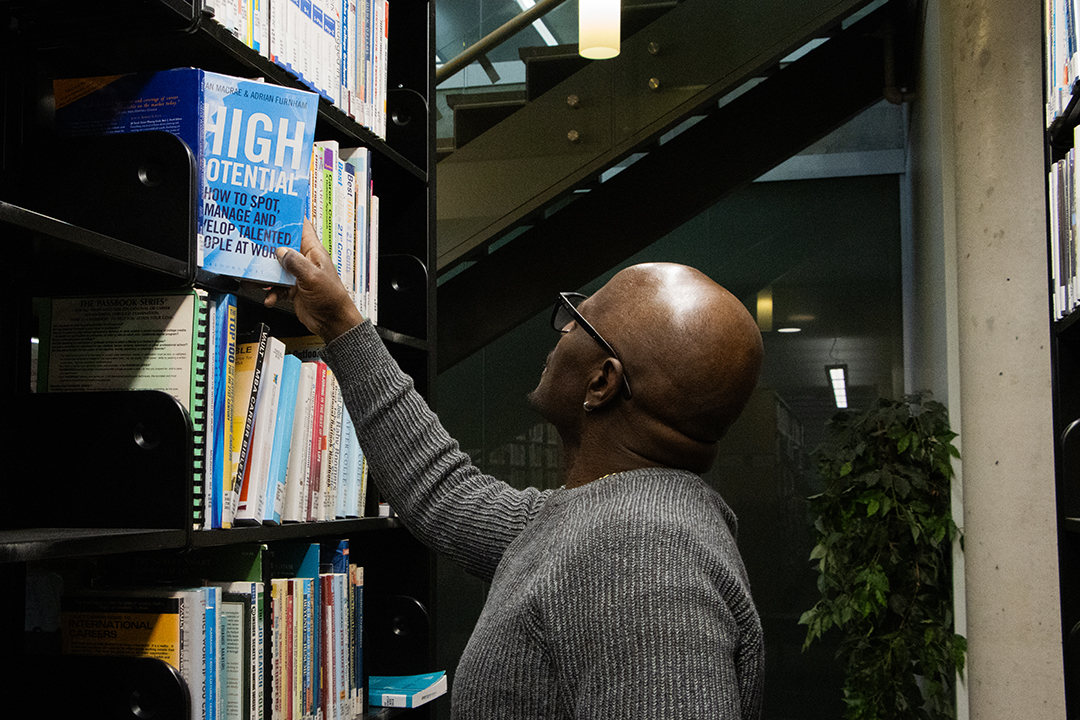 A black man wearing glasses and a grey sweater reaches up to take a blue book titled "High Potential" from a library shelf.