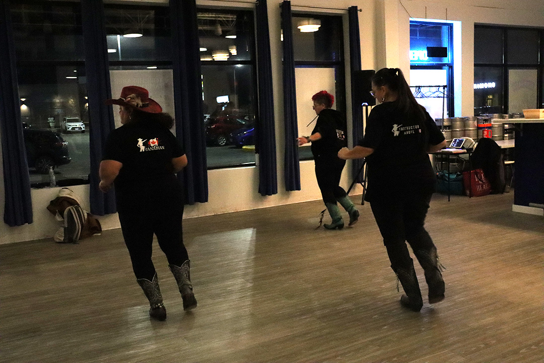 Three women wearing black pants, cowboy boots and a black shirt with red and white writing. The all dance on a wooden floor with floor to ceiling windows in front of them. The time of day in late with the sun having gone down.