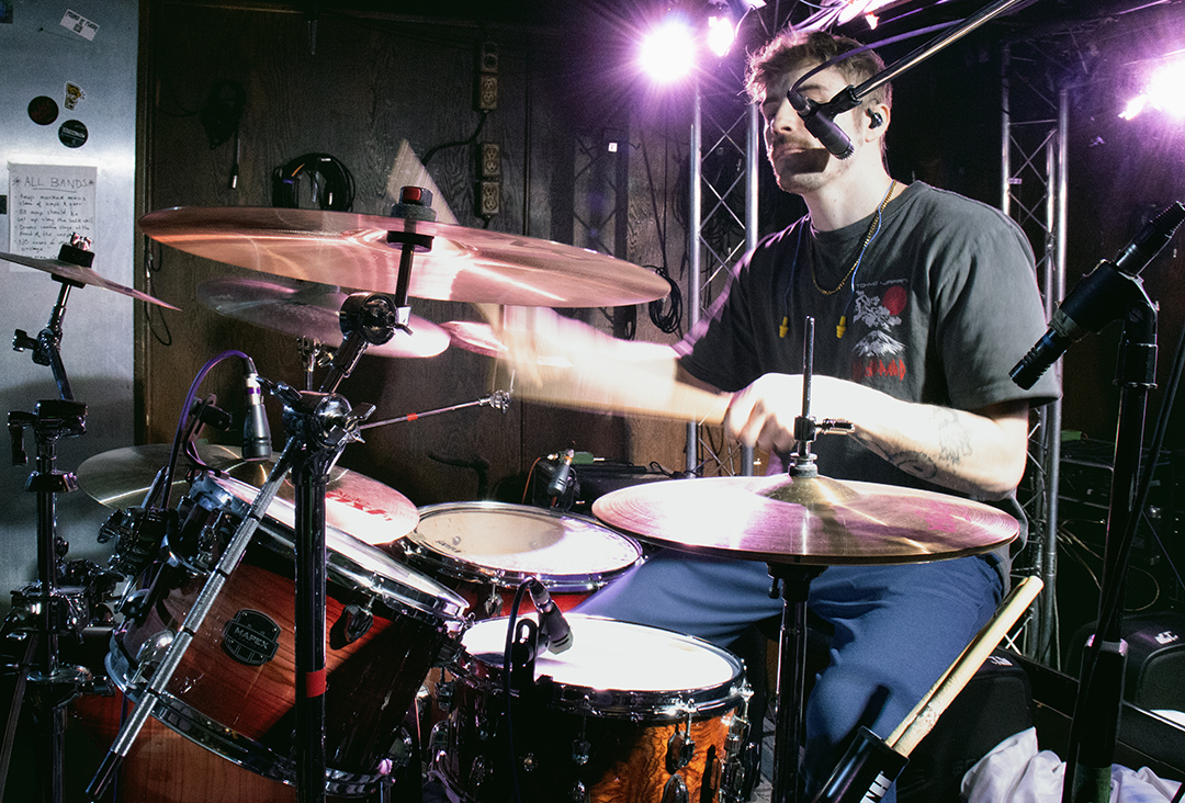 A young man plays drums on stage with lights flashing behind him and a microphone near his face.
