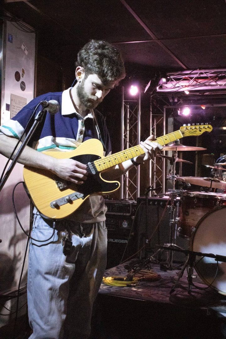 A young man wearing a t-shirt and blue jeans, holding a yellow guitar, looks down while performing on stage.