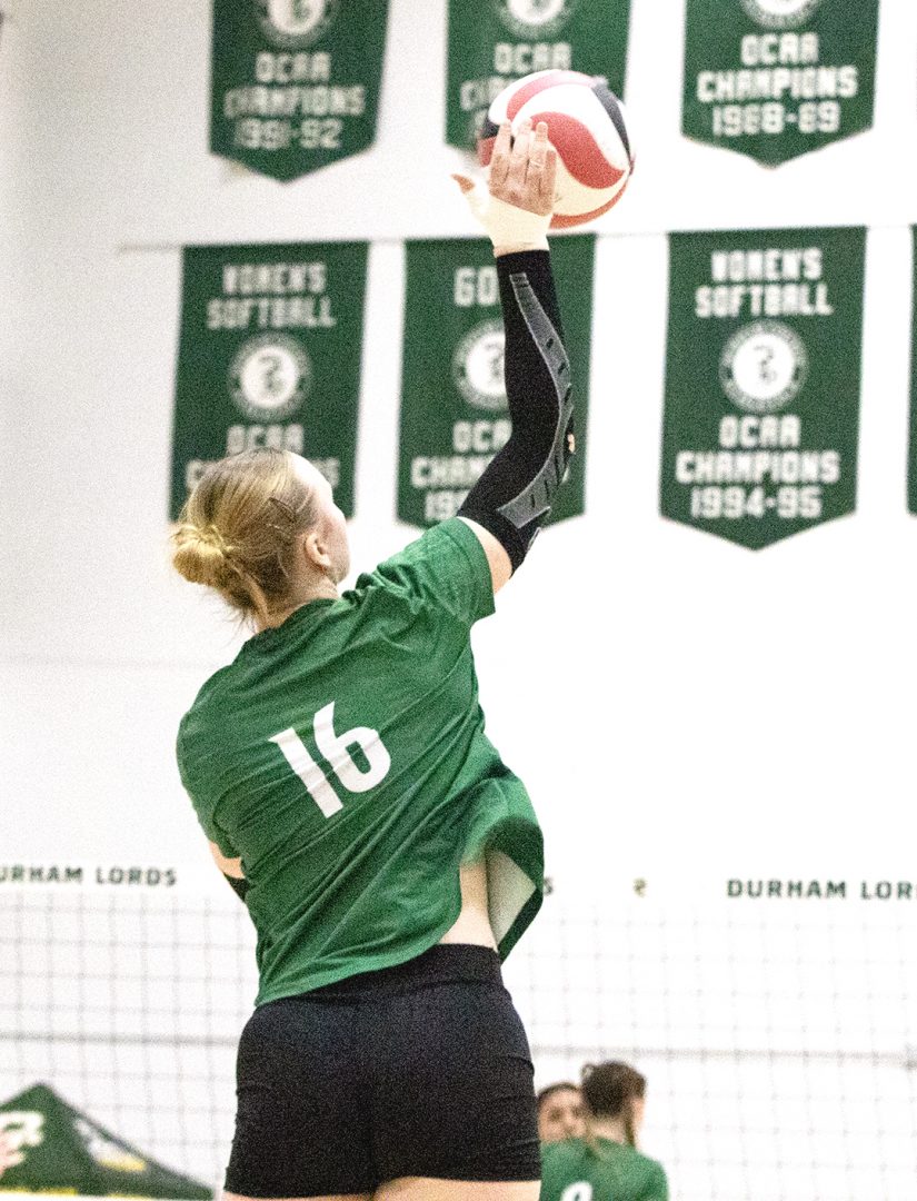 A female athlete in a green jersey with number 16 jumps to make a serve during a volleyball match