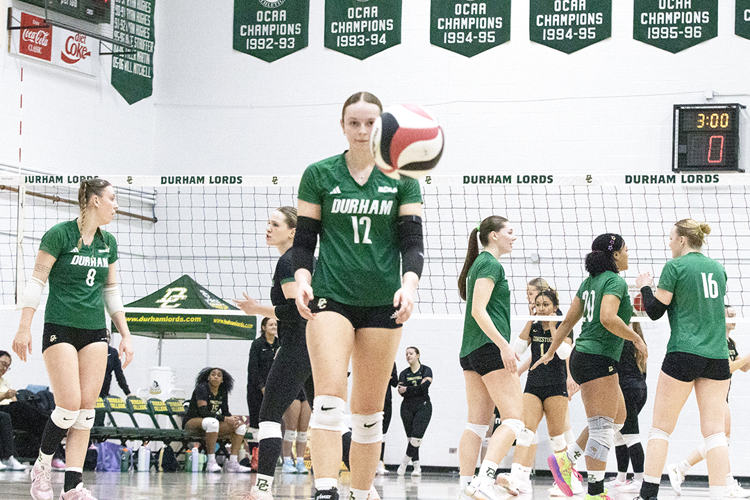 A female athlete walks towards the camera, and a volleyball is seen in the air in the foreground, with other team members in the background during a volleyball match.