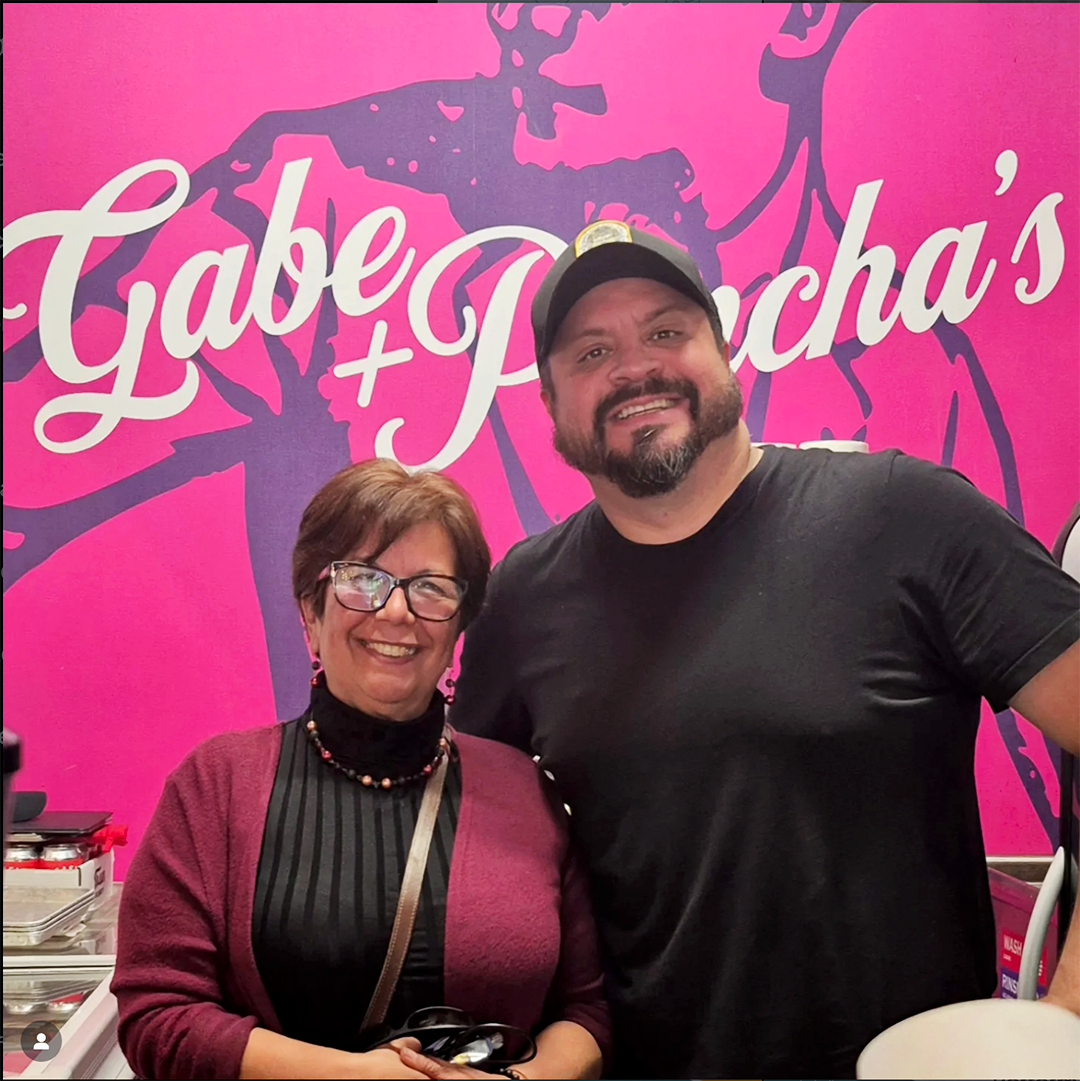 A woman on the left wearing glasses, a black turtleneck, and a red sweater. A man on the right with a beard, wearing a black T-shirt and a cap.