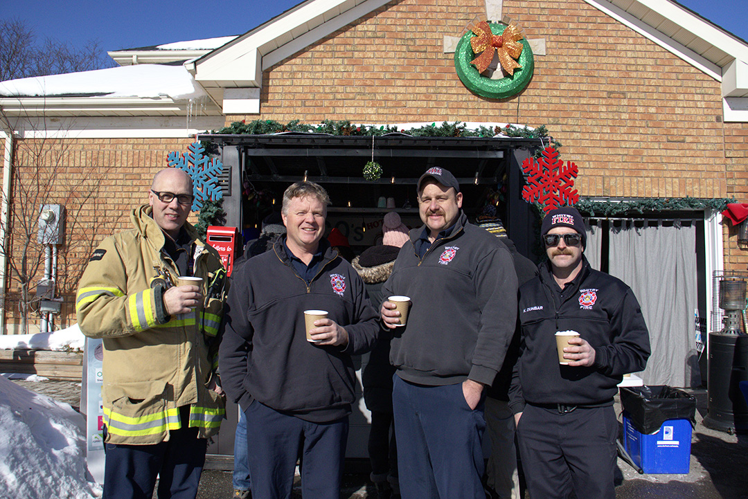 Four men stand in front of a booth. One is wearing a firefighter coat and three are wearing dark fleeces.