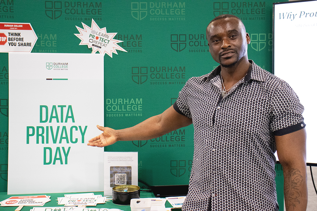 A young man in a black and white shirt points to a white sign that says Data Privacy Day in green letters.