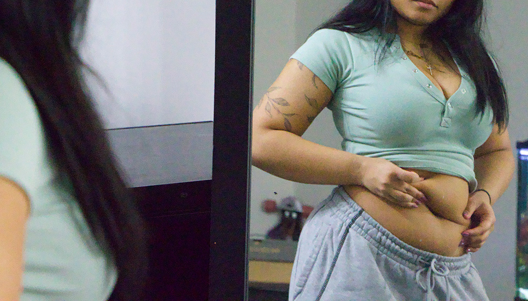 A young woman wearing grey sweatpants and a light green shirt stands in front of a mirror grabbing at her stomach.