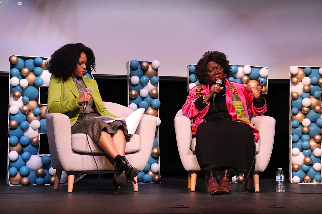 Two women holding microphones sit in chairs on a stage.