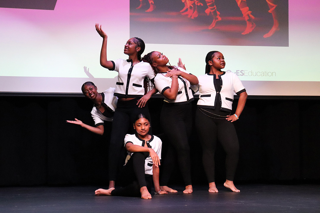 Five young women pose together on a stage. They are wearing black pants and white shirts with black trim.
