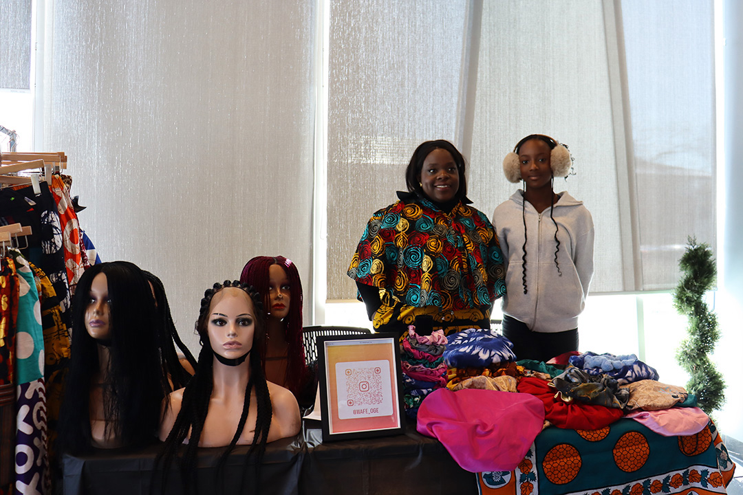 A woman and her daughter stand behind a table displaying bonnets, wigs and clothing.