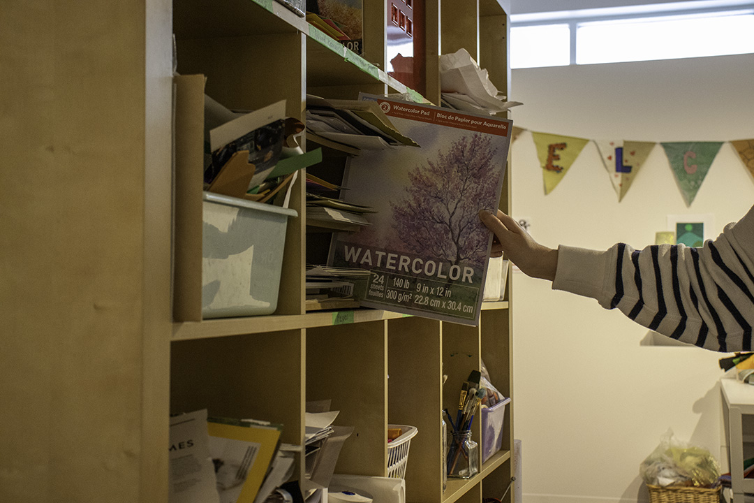 A hand pulls a book titled "Watercolor" from a bookshelf. The bookshelf is filled with art supplies.