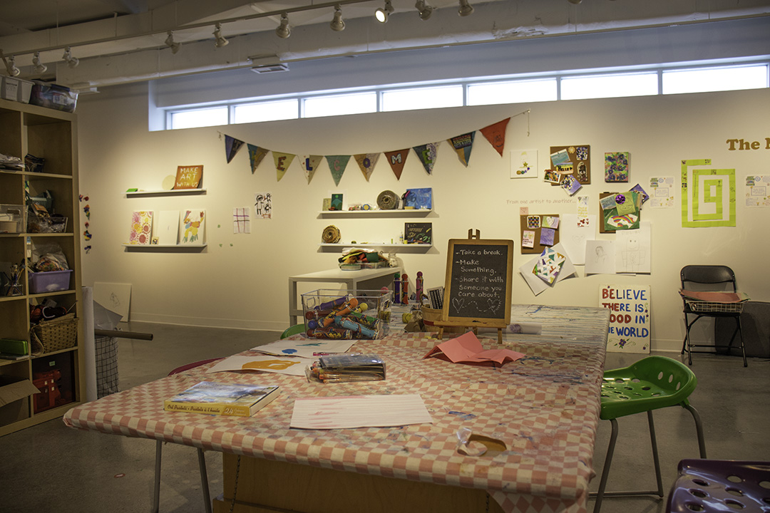 A table with a red and white checked tablecloth on top is in a large studio space. There are markers, paper and bingo dabbers on the table, and stools around it.
