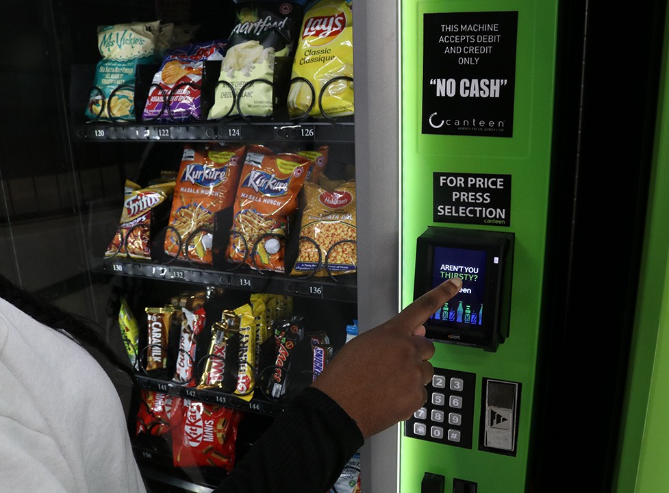 A girl wearing a black long sleeved shirt is picking a snack in a green and black vending machine with a black sign on it that says "no cash" in bold, capital letters.