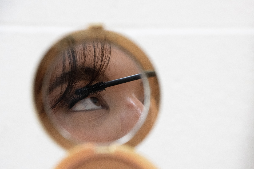 A young woman's eye is reflected in a compact mirror as she applies mascara.