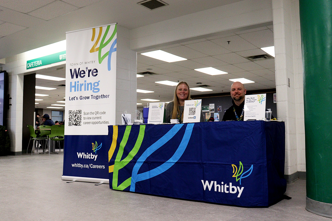 A blonde haired woman and a bald man with a beard sits behind a table draped with a dark blue tablecloth and the words "Whitby" written in white in the corner. A large sign is placed to the left of them that reads "We&squot;re Hiring" in bold lettering.